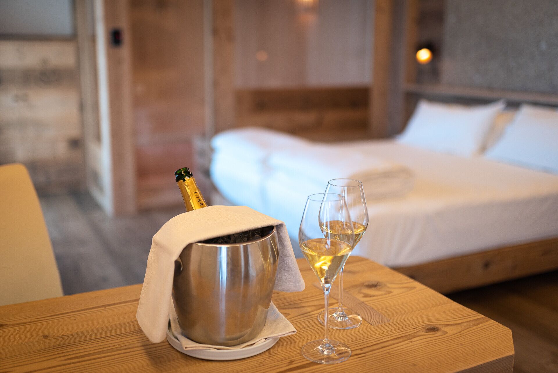 Et træbord med en champagneflaske og to champagneglas, en seng med hvide sengetøj og en trægulv.