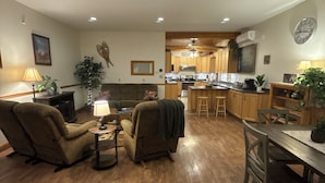 A spacious living room with a sofa, a coffee table, and a dining table with chairs. There is a kitchen area with wooden cabinets and a refrigerator. - Prime Snowmobile-Friendly Villa in Northern Maine  Winter Trails & Comfort (Bridgewater)