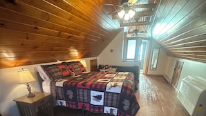 Une chambre confortable avec un plafond en bois, un lit recouvert d’une couverture à carreaux, une lampe de chevet et un tapis rond.
