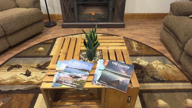 A living room with a fireplace, two sofas, a coffee table with magazines, a lamp, and a framed picture on the wall. - Prime Snowmobile-Friendly Villa in Northern Maine  Winter Trails & Comfort (Bridgewater)