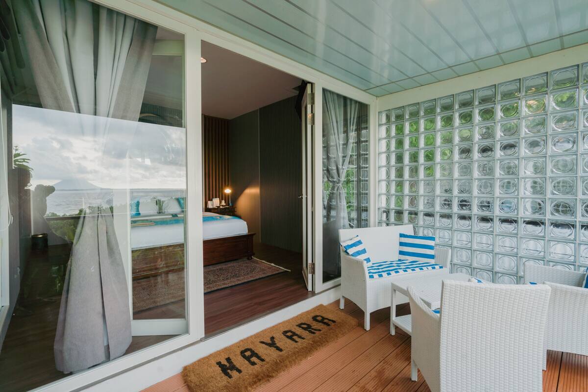 Una habitación de hotel con una puerta corrediza de vidrio que da a un balcón, una cama, una pequeña mesa con sillas y una pared de bloques de vidrio.