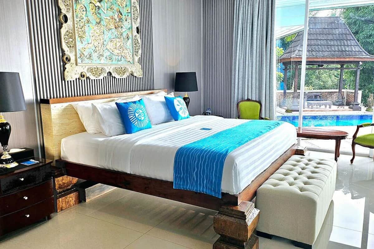 Un dormitorio amplio con una cama grande, cabecera de madera, mesita de noche con lámpara, sillón verde y vistas a una piscina y un pabellón a través de amplios ventanales.