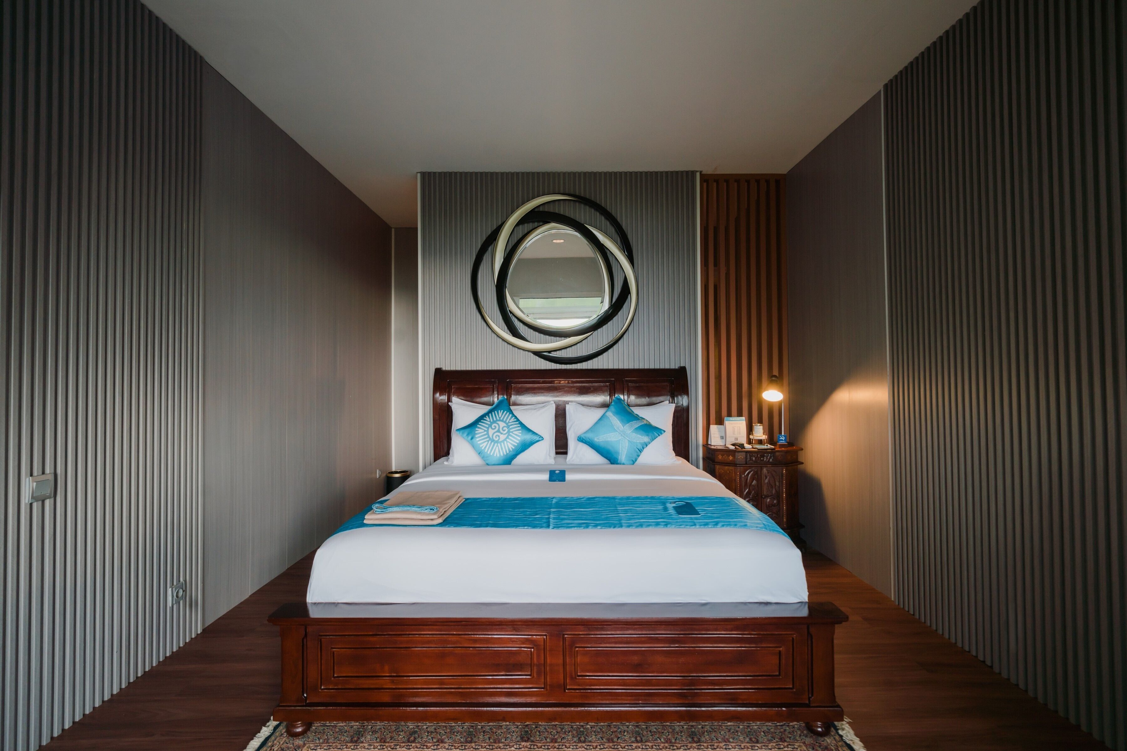 Una cama bien hecha con cabecera de madera, una mesita de noche con lámpara y un espejo fijado a la pared.