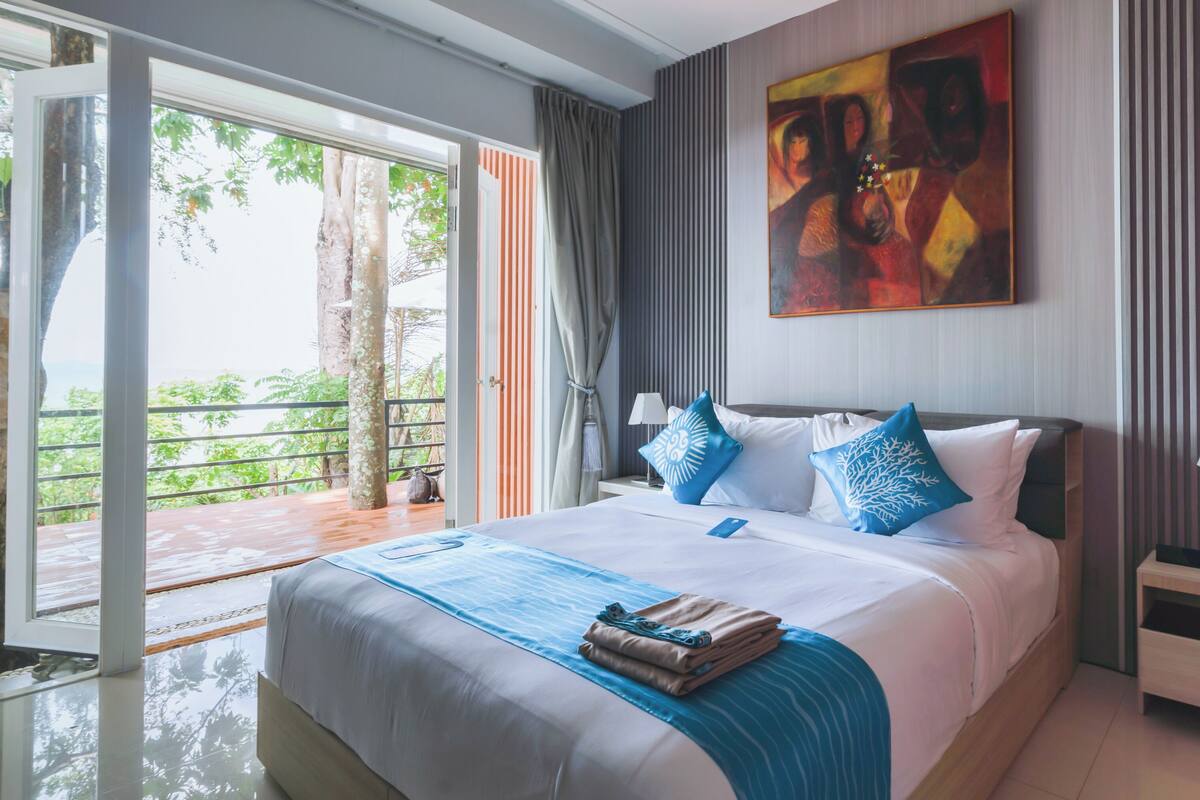 Habitación de hotel con cama, mesita de noche, un cuadro y vistas al exterior con zonas verdes.