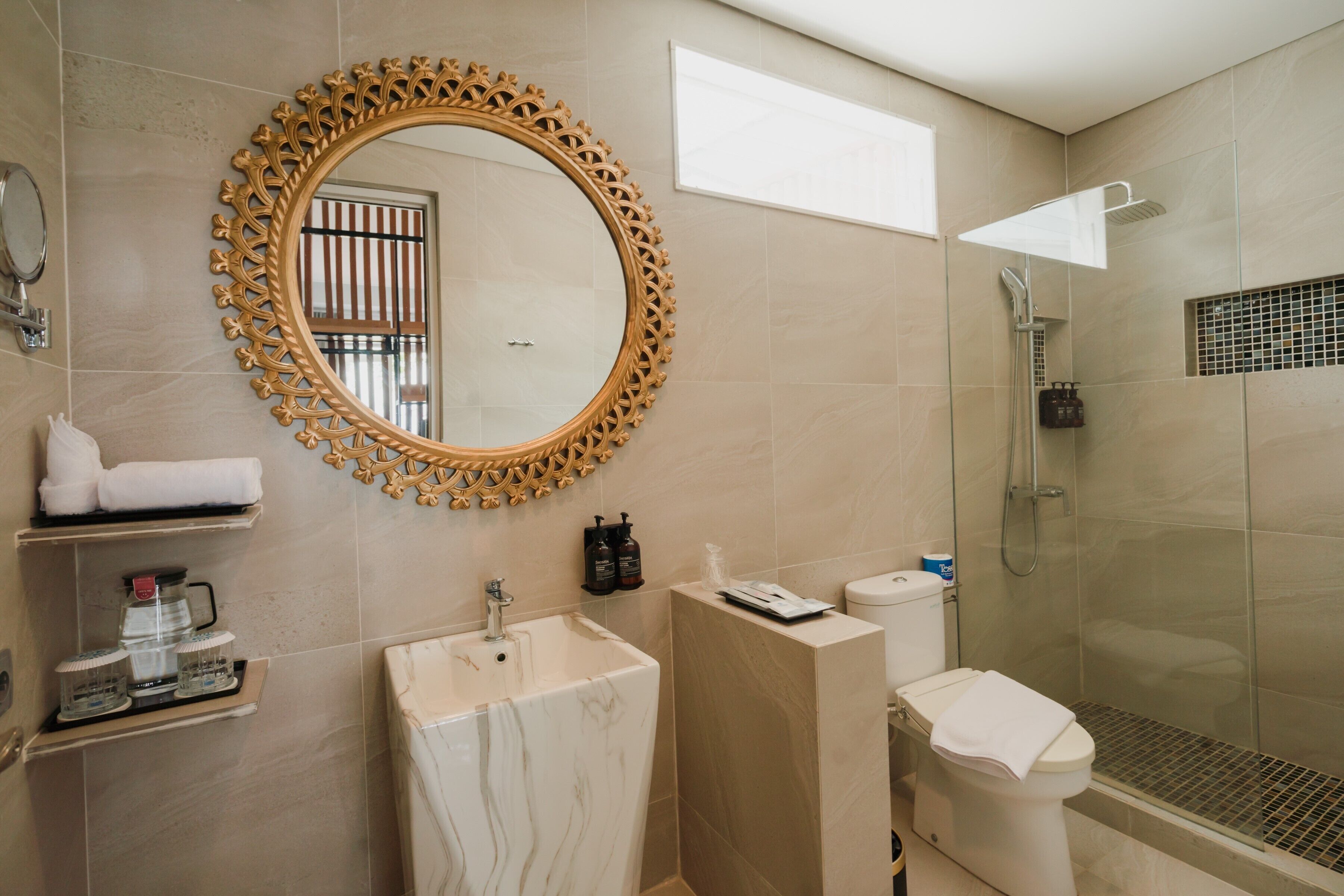 A modern bathroom with a marble sink, a round mirror with a decorative frame, a glass-enclosed shower, and a toilet.