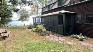 A two-story house with a covered porch, a small garden, and a view of the water. - LARGE RUSTIC COTTAGE WITH BUNK HOUSE.  SLEEPS 17.  (Sebago)