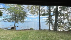 A view from inside a cabin with a window overlooking a grassy area with trees and a body of water. - LARGE RUSTIC COTTAGE WITH BUNK HOUSE.  SLEEPS 17.  (Sebago)