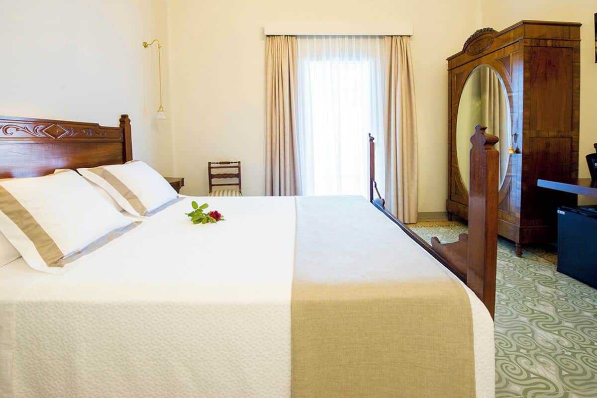 Una cama bien hecha con colcha blanca y beige, cabecera de madera y una pequeña rosa sobre la cama. Hay un armario de madera con espejo, un escritorio y una silla en la habitación.