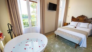A hotel room with a large oval bathtub filled with water and rose petals, a bed with a wooden headboard, and a balcony with a view of greenery. - Bolet Casa Modernista (Castellvi de la Marca)