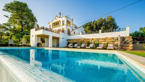 A poolside view of a modern building with a stone wall and a white facade, featuring a pool with clear blue water and lounge chairs. - Bolet Casa Modernista (Castellvi de la Marca)