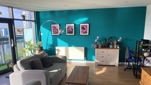 Ein modernes Wohnzimmer mit einem grauen Sofa, einem Holztisch und einer türkisblauen Akzentwand. An den Wänden hängen gerahmte Bilder, ein weißes Schränkchen und ein Schreibtisch mit Stuhl.