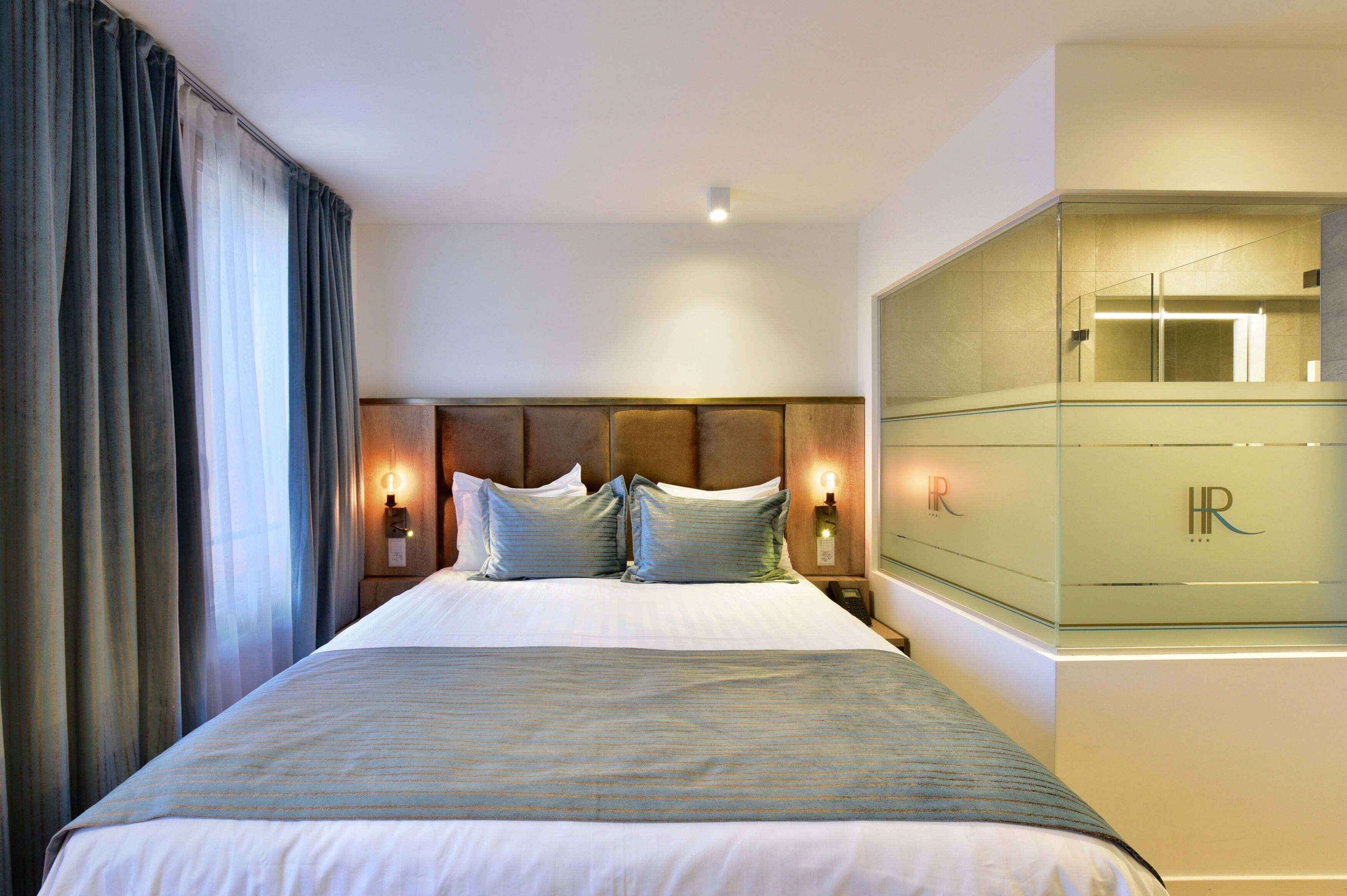 A hotel room with a large bed, a nightstand with a lamp, and a glass cabinet with a mirror.