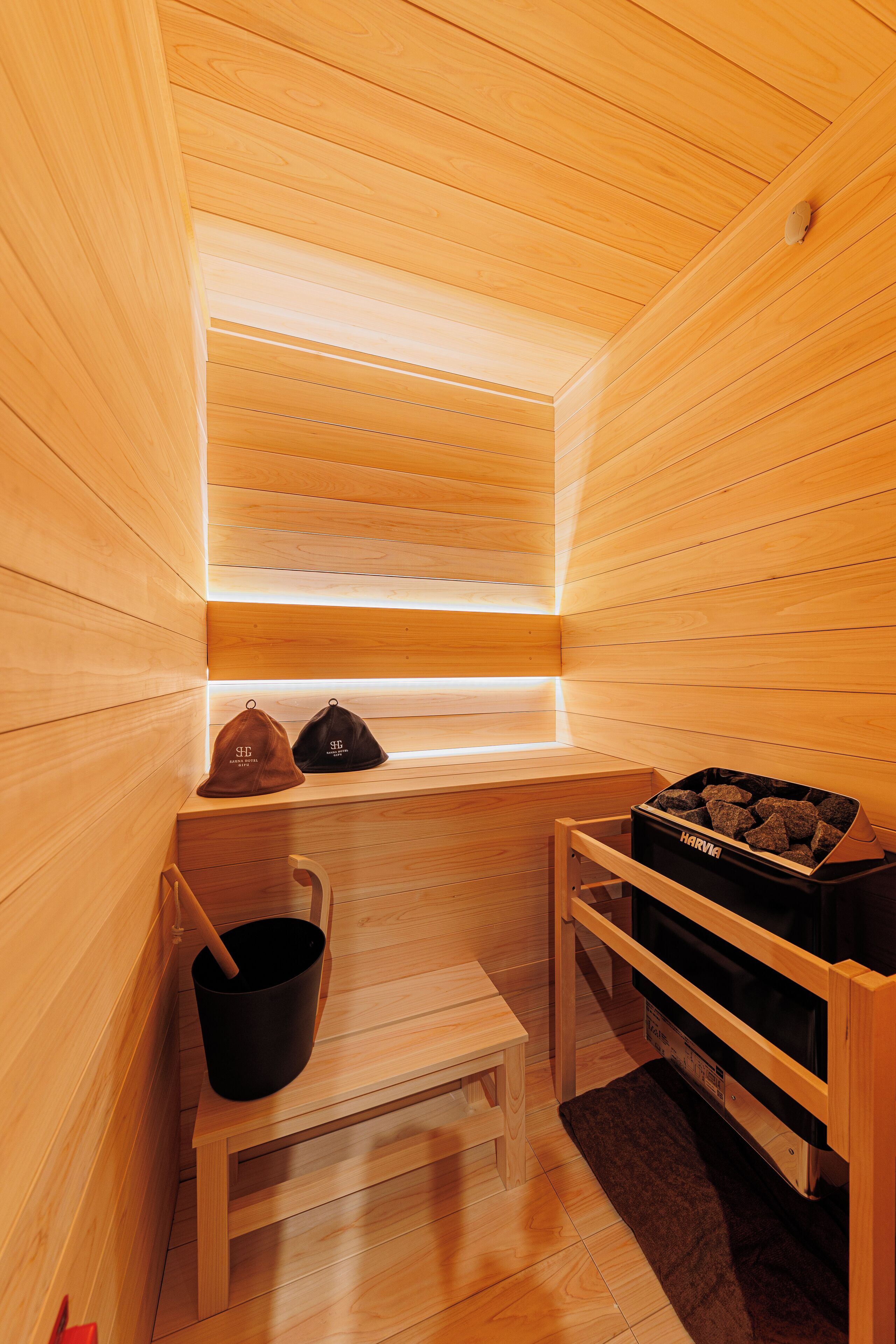 A sauna with wooden walls and a bench. There are two hats on the bench and a bucket.