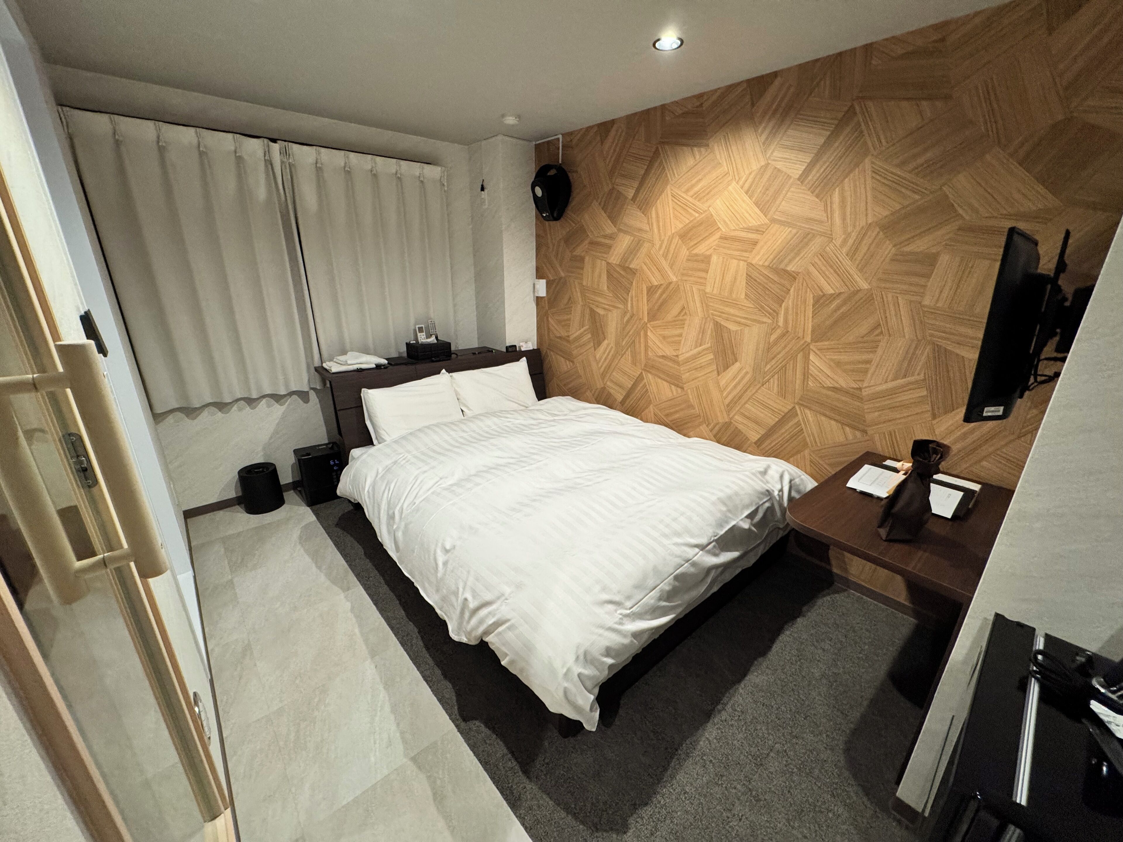 A modern hotel room with a large bed, a wooden accent wall, a bedside table with a lamp, and a small desk with a chair.