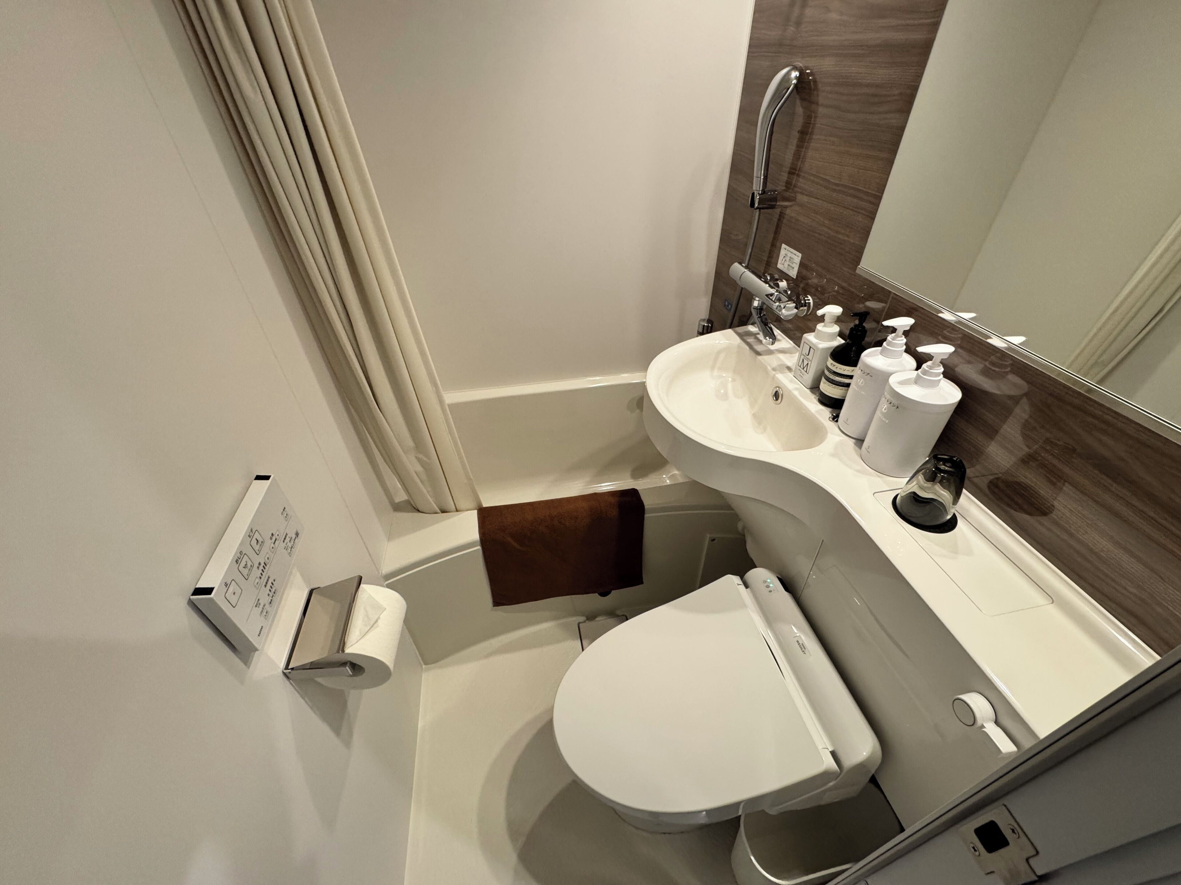 A modern bathroom with a white sink, toilet, and shower. There is a towel rack with a brown towel, a mirror, and toiletries on the sink.