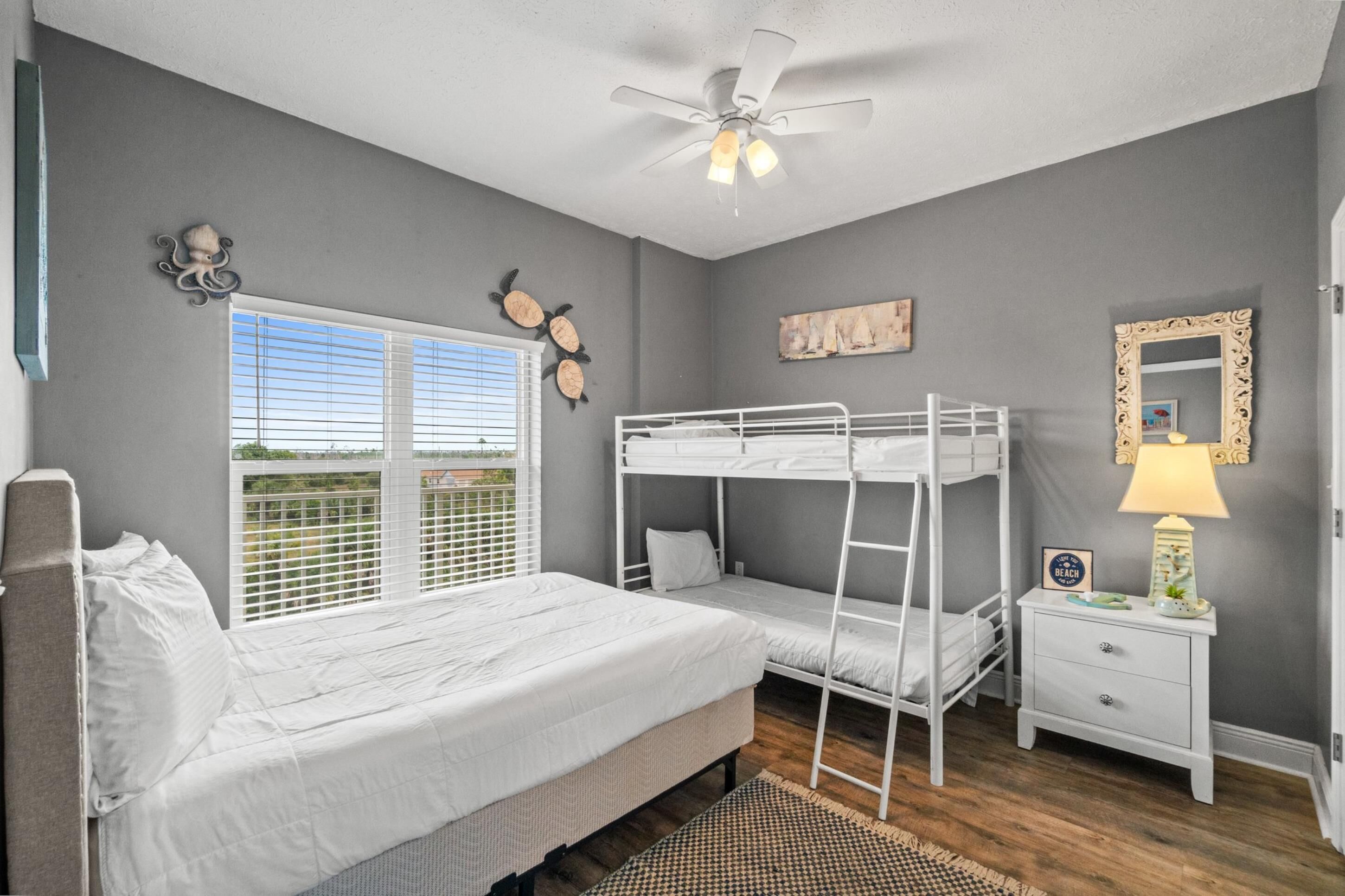 A bedroom with a bunk bed, a single bed, a nightstand with a lamp, a ceiling fan, and a window with blinds.