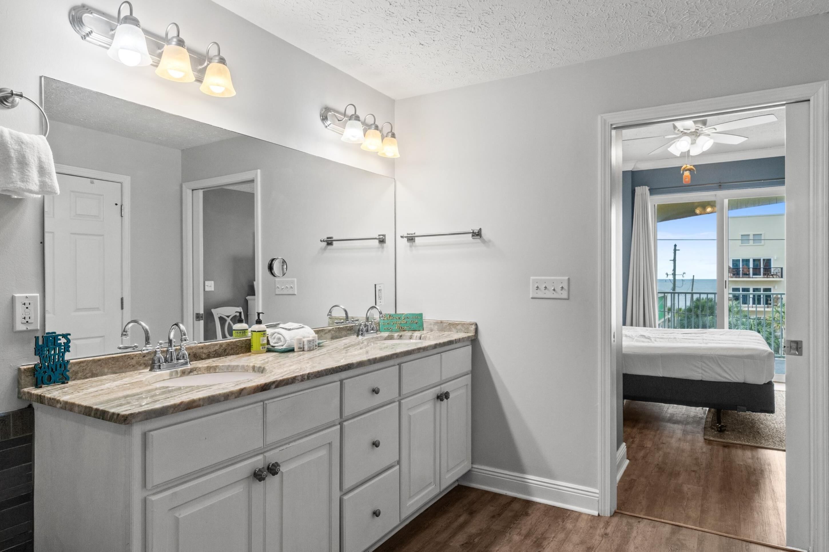 A bathroom with a large mirror, a sink with a marble countertop, and a vanity with drawers. There is a towel rack on the wall and a ceiling fan in the corner.