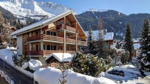 A wooden chalet with a balcony, surrounded by snow-covered trees and mountains. - Luxurious Alpine Retreat in Verbier Place Centrale with Panoramic Mountain Views (Val de Bagnes)