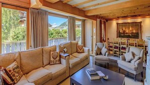 A cozy living room with a sofa, armchairs, and a coffee table. There is a large window with curtains, a painting on the wall, and a bookshelf with books. - Luxurious Alpine Retreat in Verbier Place Centrale with Panoramic Mountain Views (Val de Bagnes)