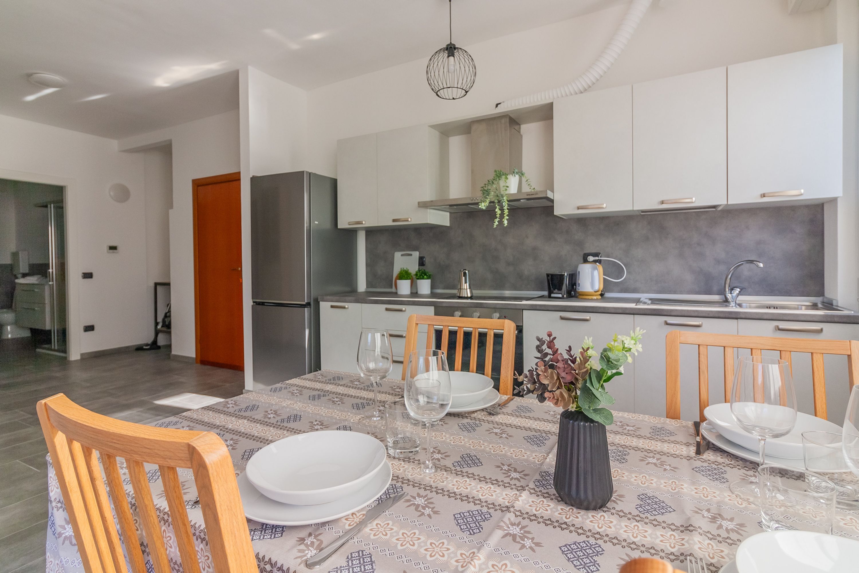 Una cocina moderna con armarios blancos, un refrigerador de acero inoxidable y un comedor con mantel estampado y sillas de madera.