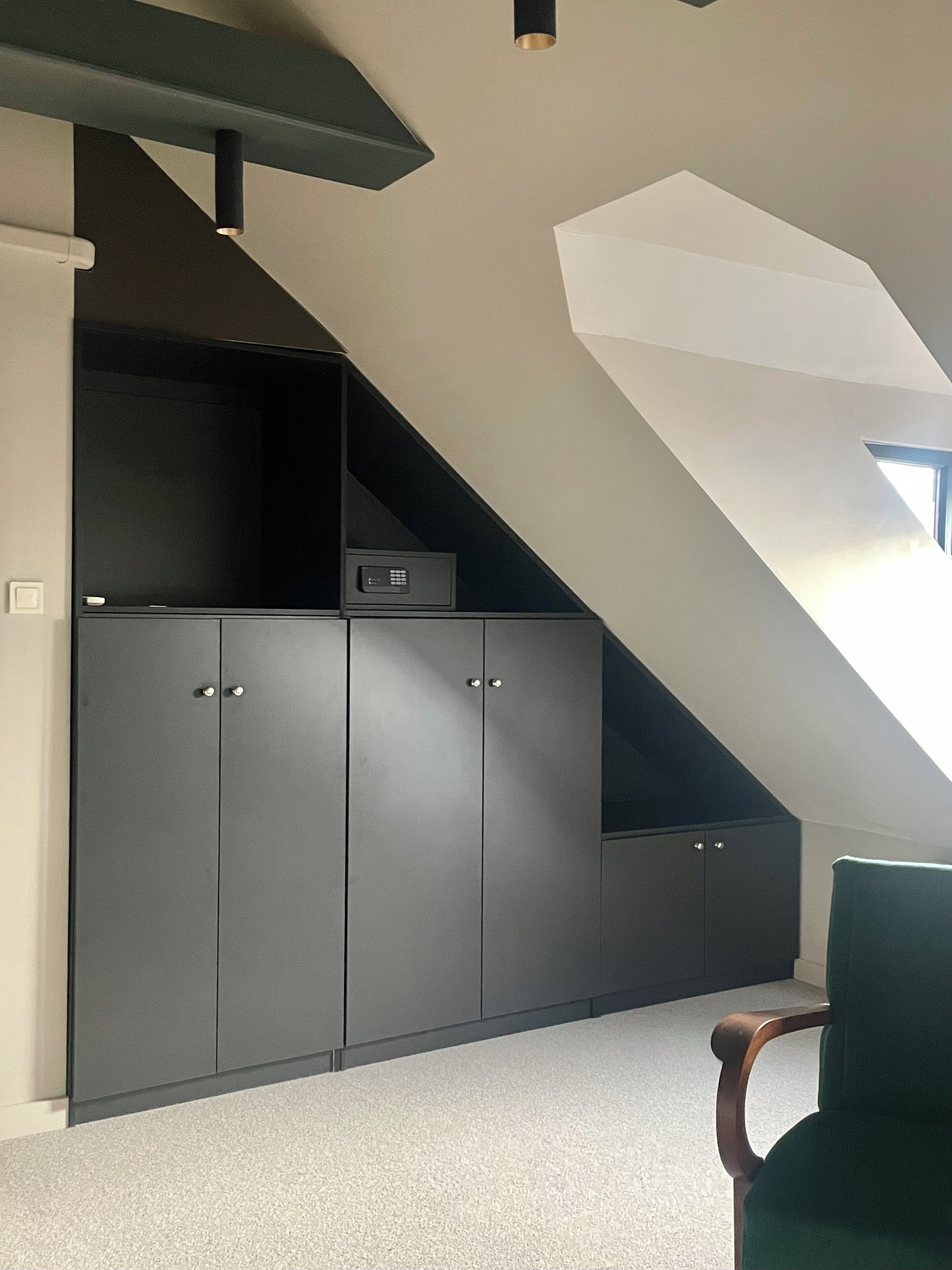 Een kamer met een schuine kap, donkergrijze inbouwmeubels en een groene stoel.