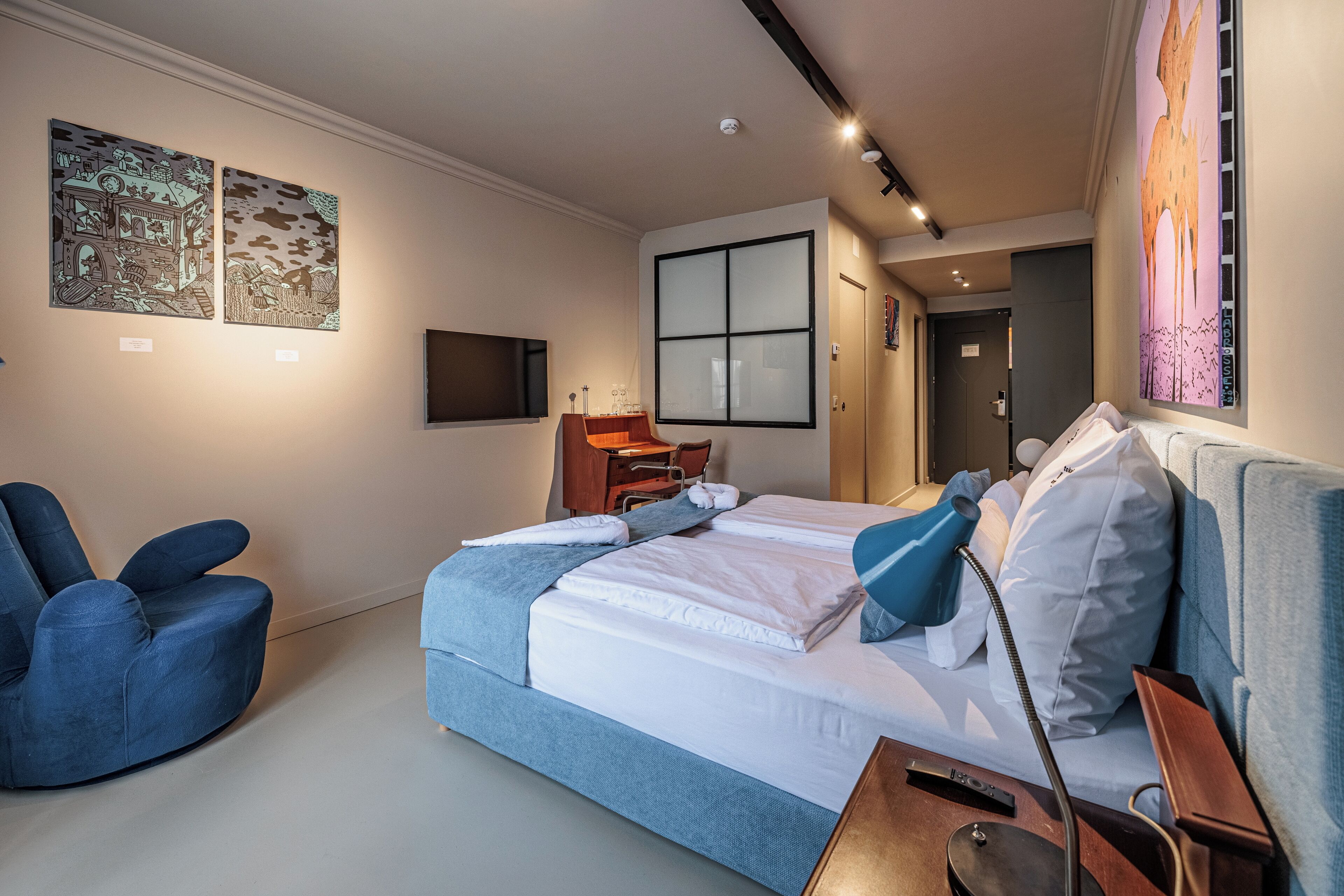 Una habitación de hotel moderna con una cama grande, un sillón azul, un escritorio y obras de arte en las paredes.