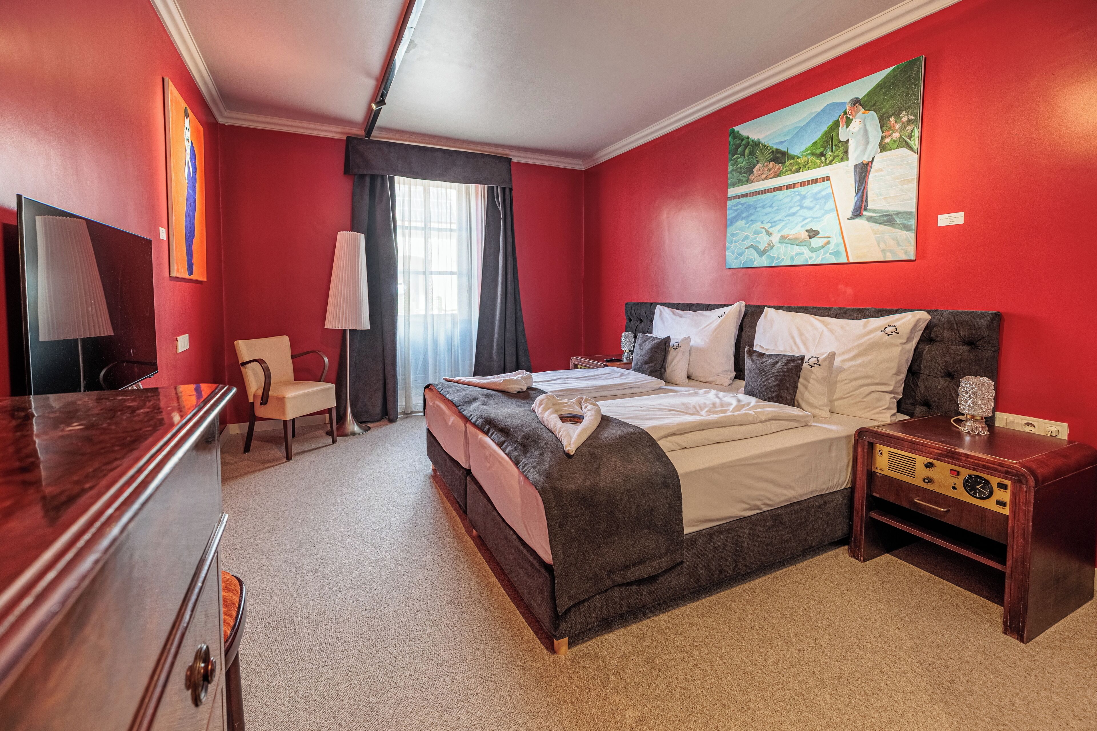 Habitación de hotel con una cama grande, una mesita de noche con radio, una silla, un televisor y un cuadro en la pared.