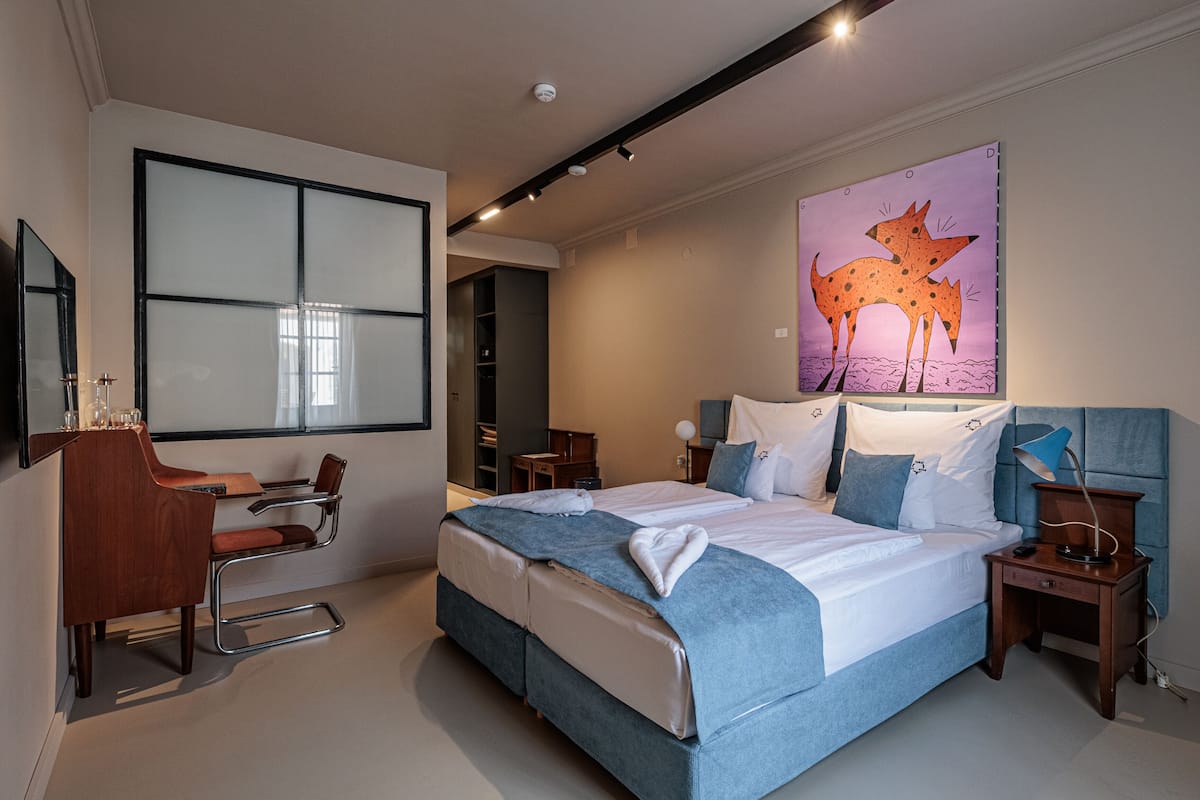 A modern hotel room with a large bed, a desk, and a chair. There is a painting on the wall and a wardrobe.
