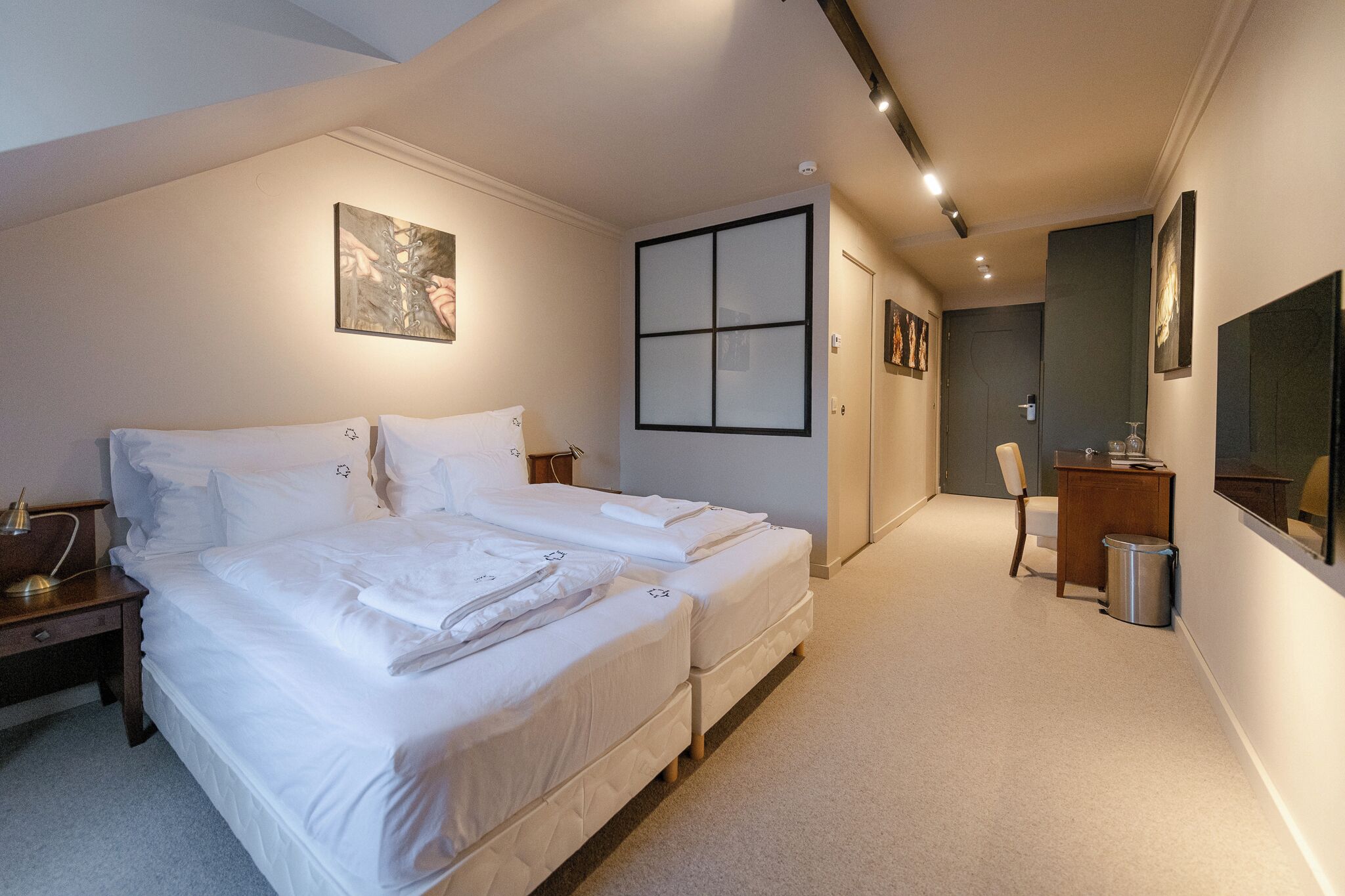 A hotel room with two beds, a desk, a chair, a trash bin, and artwork on the walls.