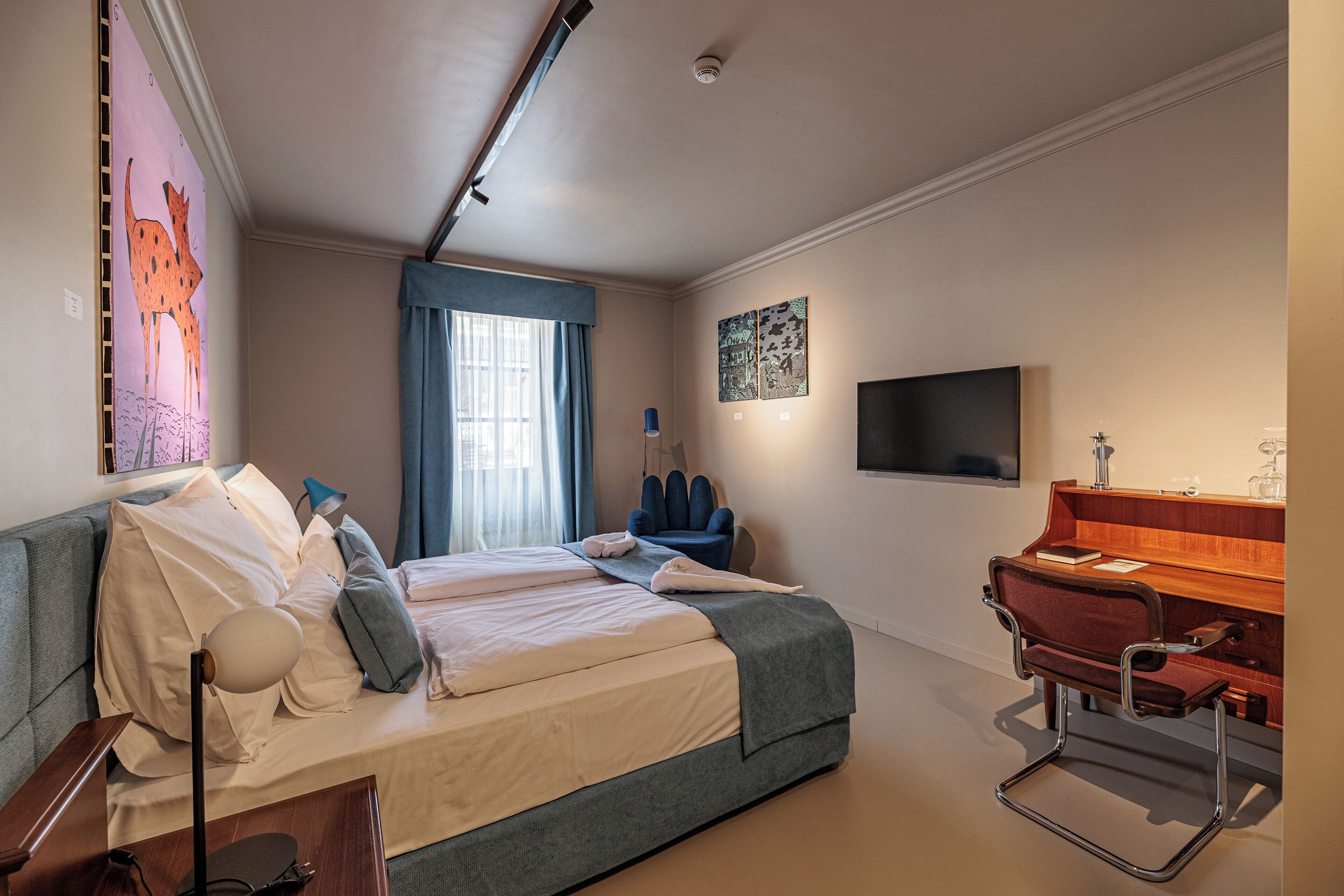 A modern hotel room with a bed, a desk, a chair, a lamp, and a TV.