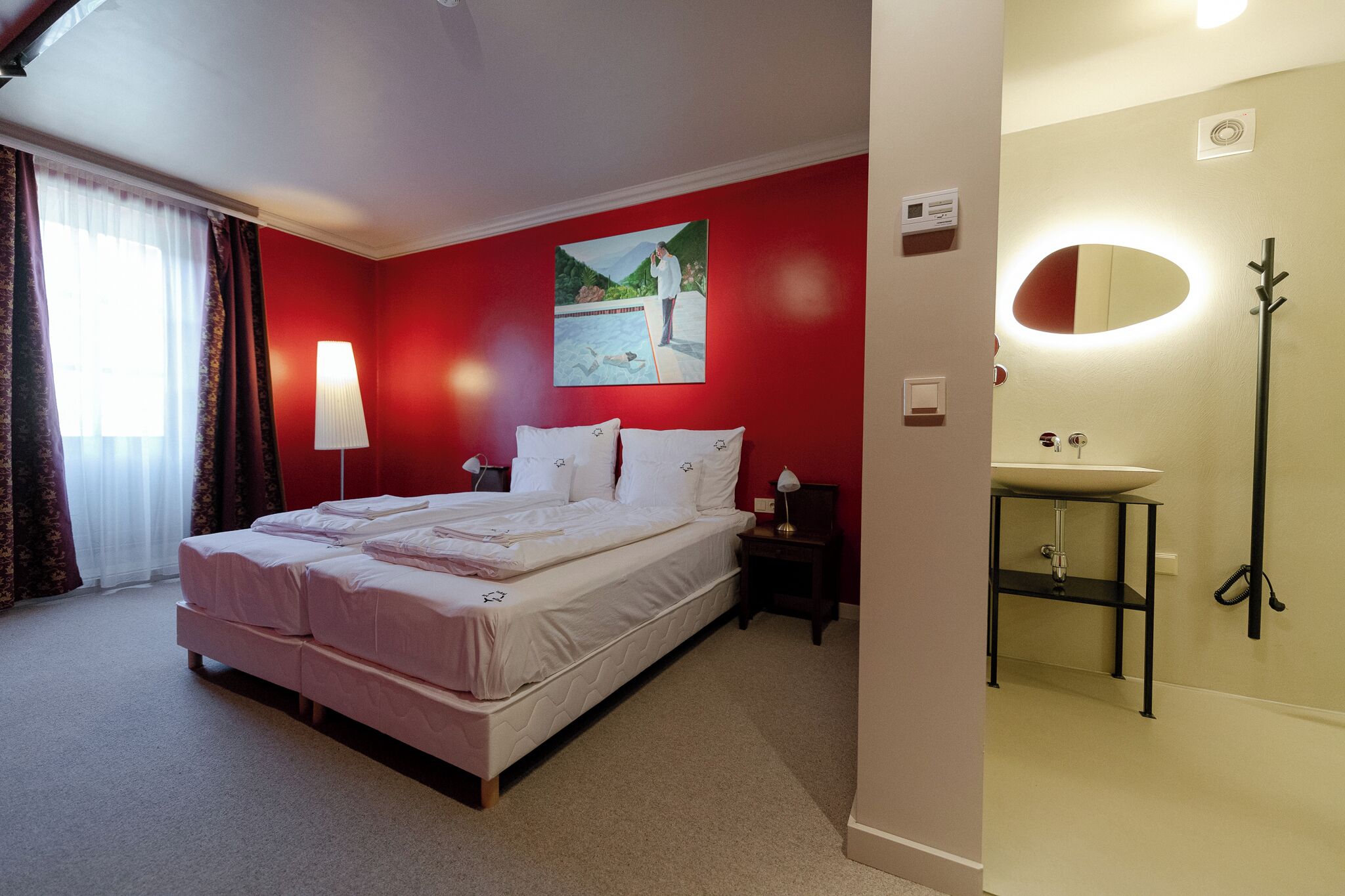 Hotelkamer met een bed, nachtkastje, lamp, schilderij, spiegel en een badkamer met wastafel en spiegel.