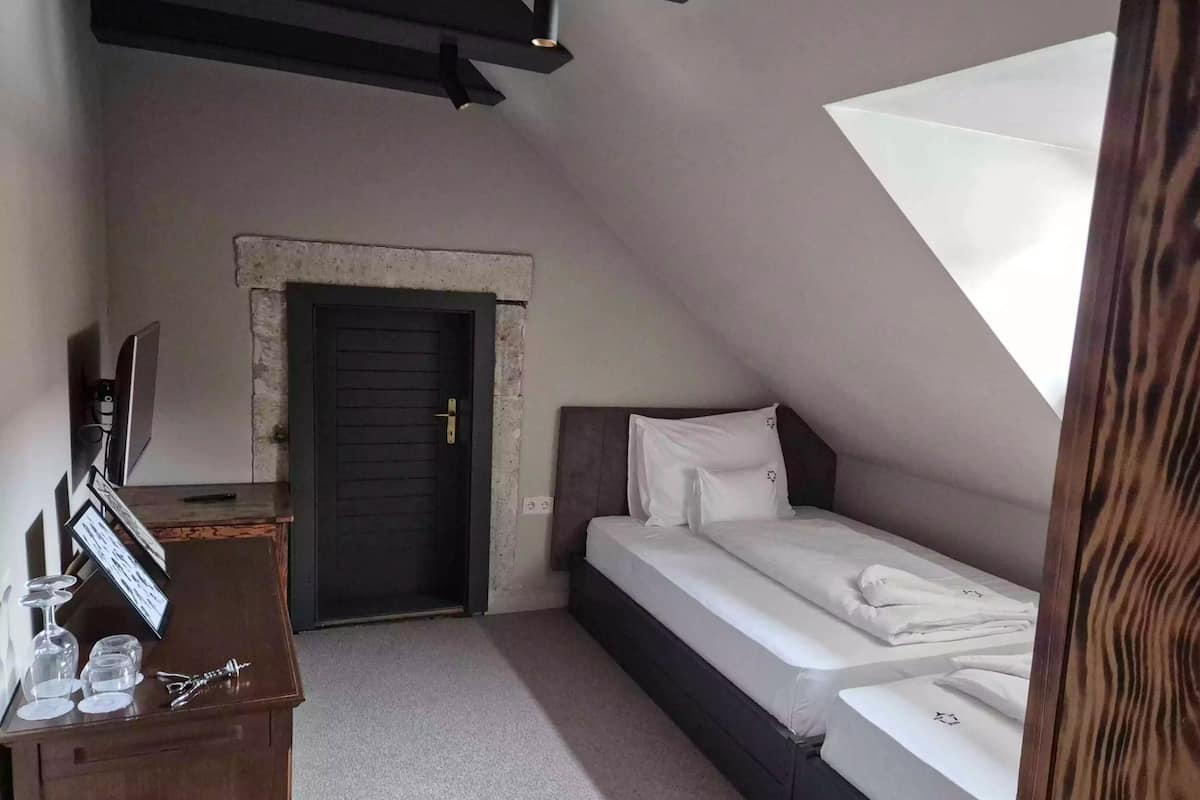 A small, dimly lit room with a single bed, a dark door, and a small table with glasses and a bottle.