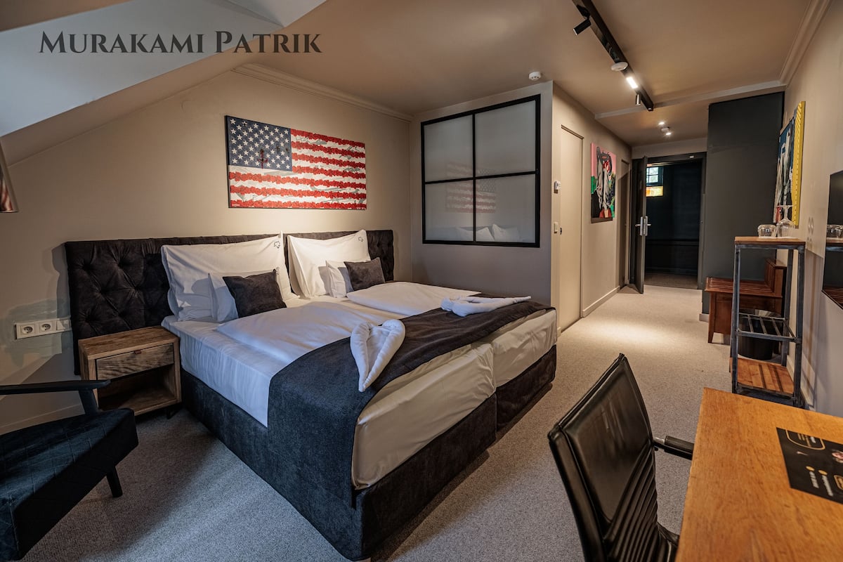 A bedroom with a large bed, a nightstand, a wooden table, and a chair. An American flag is displayed on the wall.