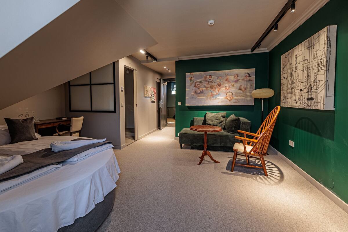 A modern hotel room with a loft bed, a small seating area with a table, and a green accent wall.