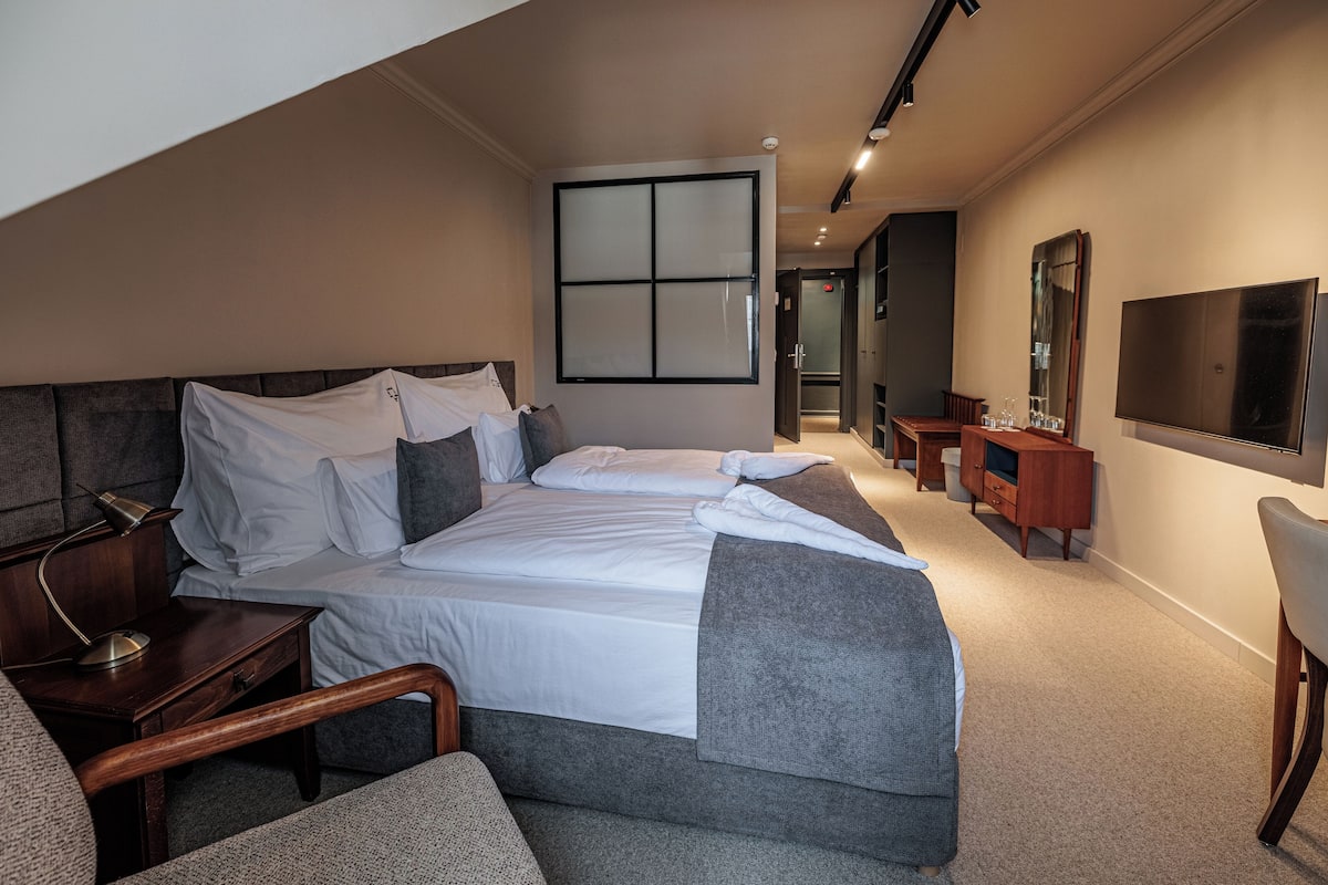 A modern hotel room with a large bed, bedside tables, a desk, a chair, a TV, and a mirror.