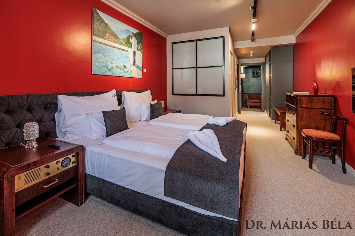 A hotel room with a large bed, a bedside table with a radio, a painting on the wall, and a view of the hallway.
