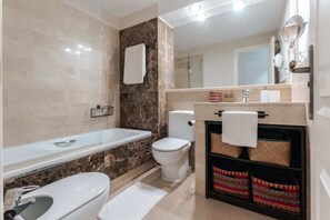 A modern bathroom with a bathtub, toilet, and sink. There are towels hanging on the wall and a shelf with baskets. - Malta by BH Marbella (Marbella)