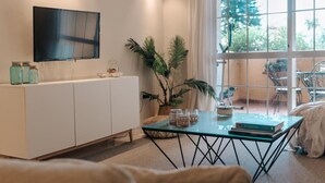 A modern living room with a glass coffee table, a white sofa, a TV mounted on the wall, and a large window with a view of greenery. - Malta by BH Marbella (Marbella)