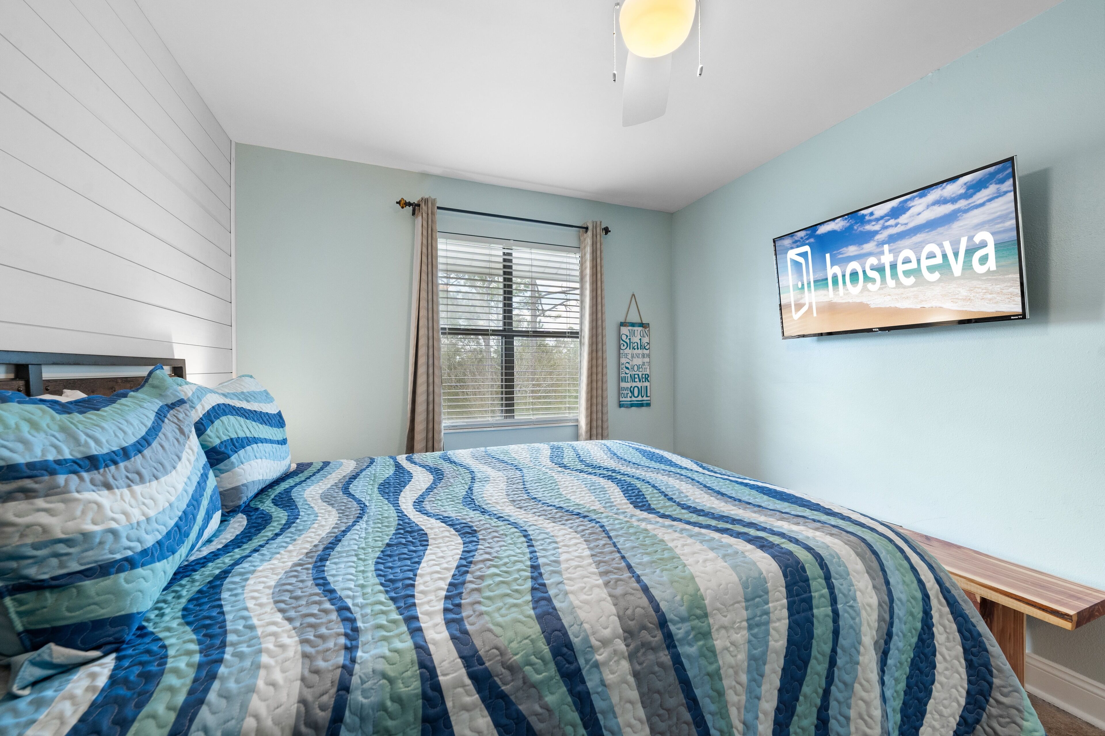 A bedroom with a bed, a TV mounted on the wall, a window with curtains, and a sign on the wall.