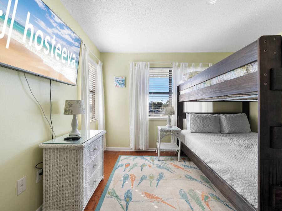 A room with a bunk bed, a desk with a lamp, a wicker dresser, and a wall-mounted TV displaying 'Hosteva'.