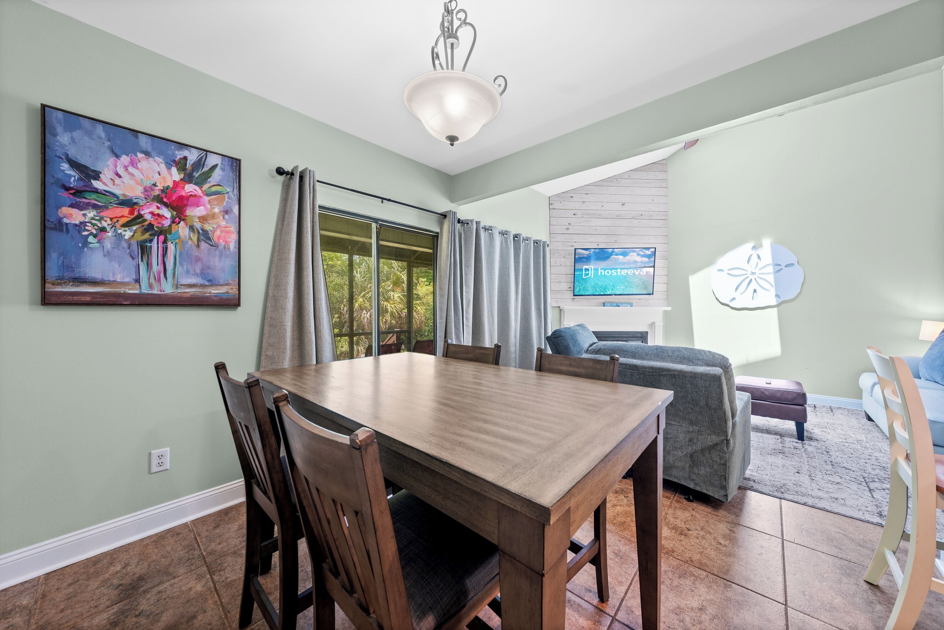 A dining area with a wooden table and chairs, a painting on the wall, and a TV in the background.