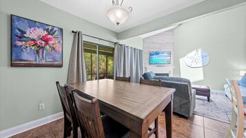 A dining area with a wooden table and chairs, a painting on the wall, and a TV in the background.