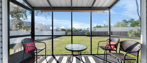 A covered outdoor area with a view of a grassy field and buildings. It includes a table and two chairs with cushions.