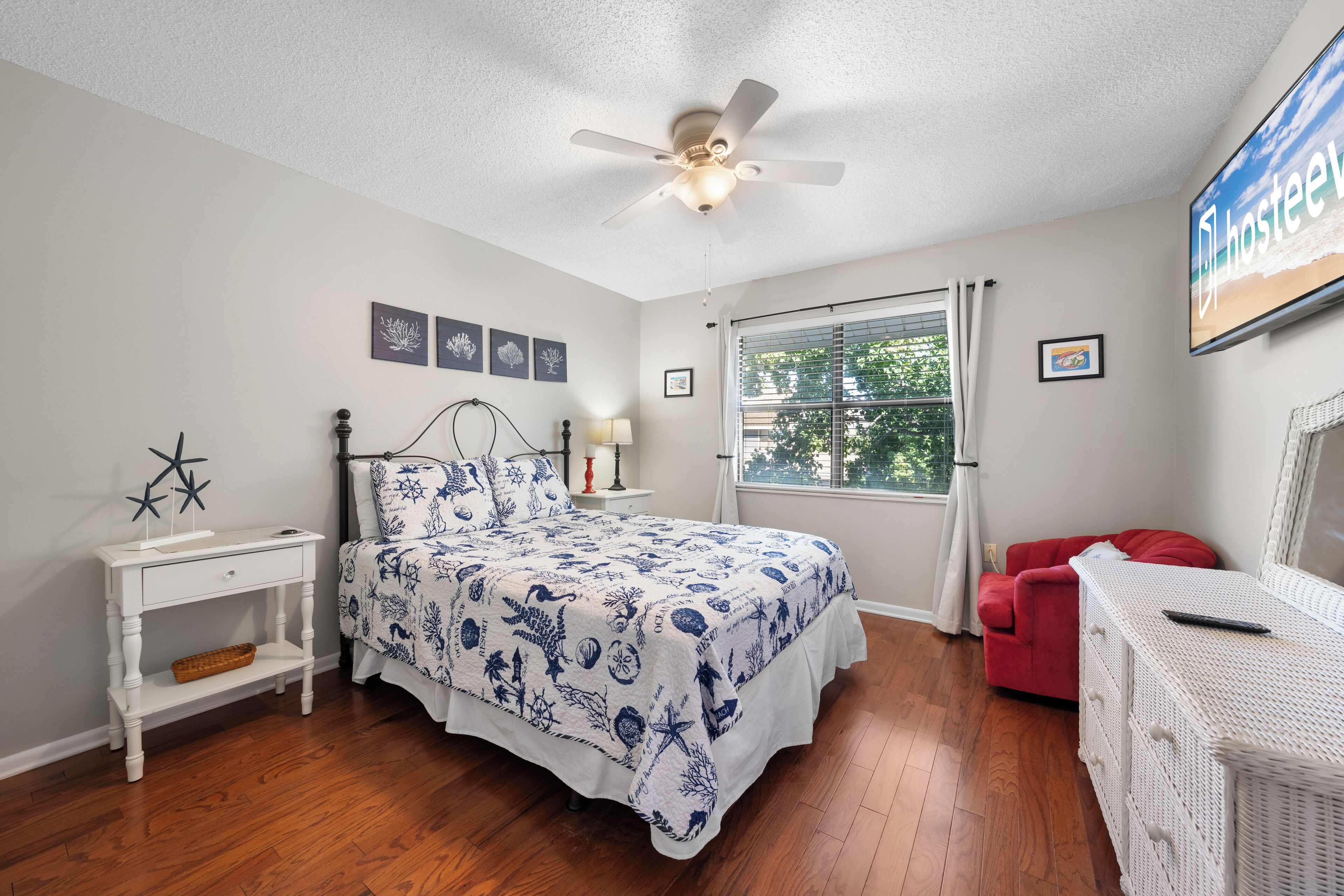 A bedroom with a bed, a nightstand, a red chair, a dresser, a ceiling fan, and a window with curtains.