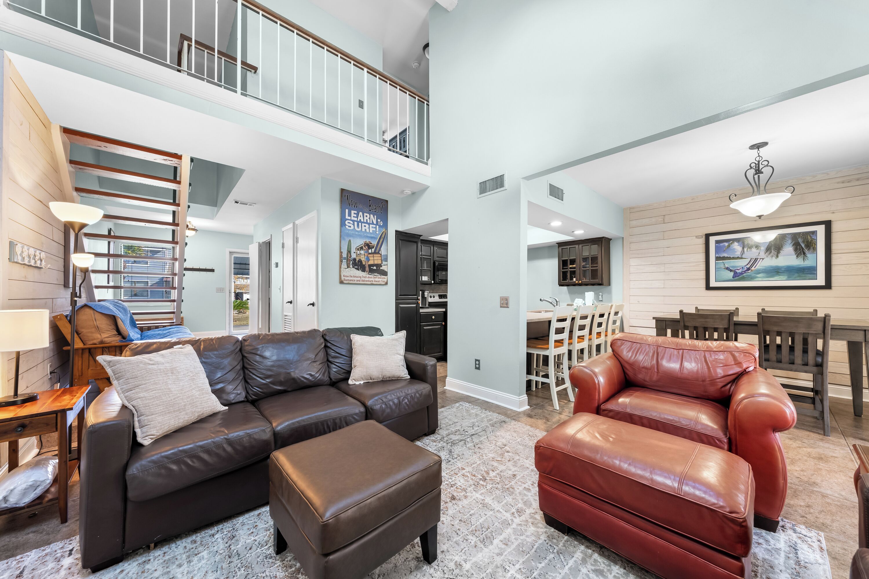 A spacious living area with a brown leather sofa, a red leather chair, and a dining table with chairs. A staircase leads to an upper level.