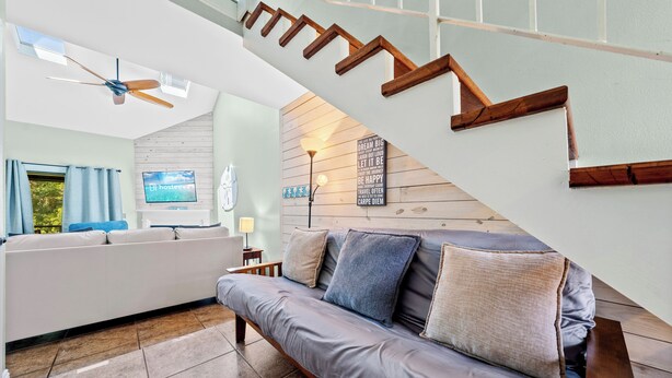Ein modernes Wohnzimmer mit grauem Sofa, einer Holztreppe und einem an der Wand befestigten Fernseher.