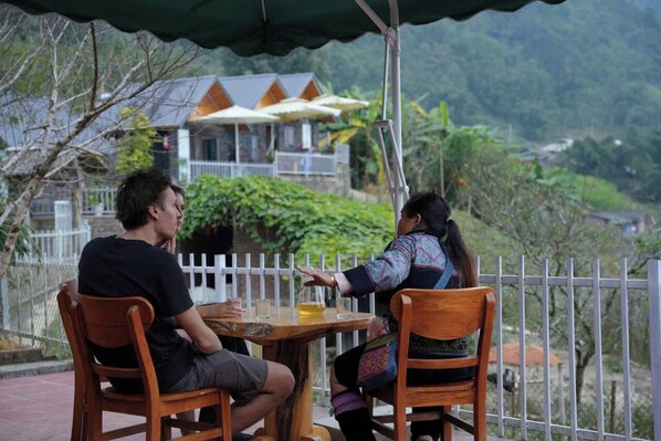 Two people sitting at a wooden table with a view of a house and greenery in the background. - Mama Lili's Homestay (Ta Van)