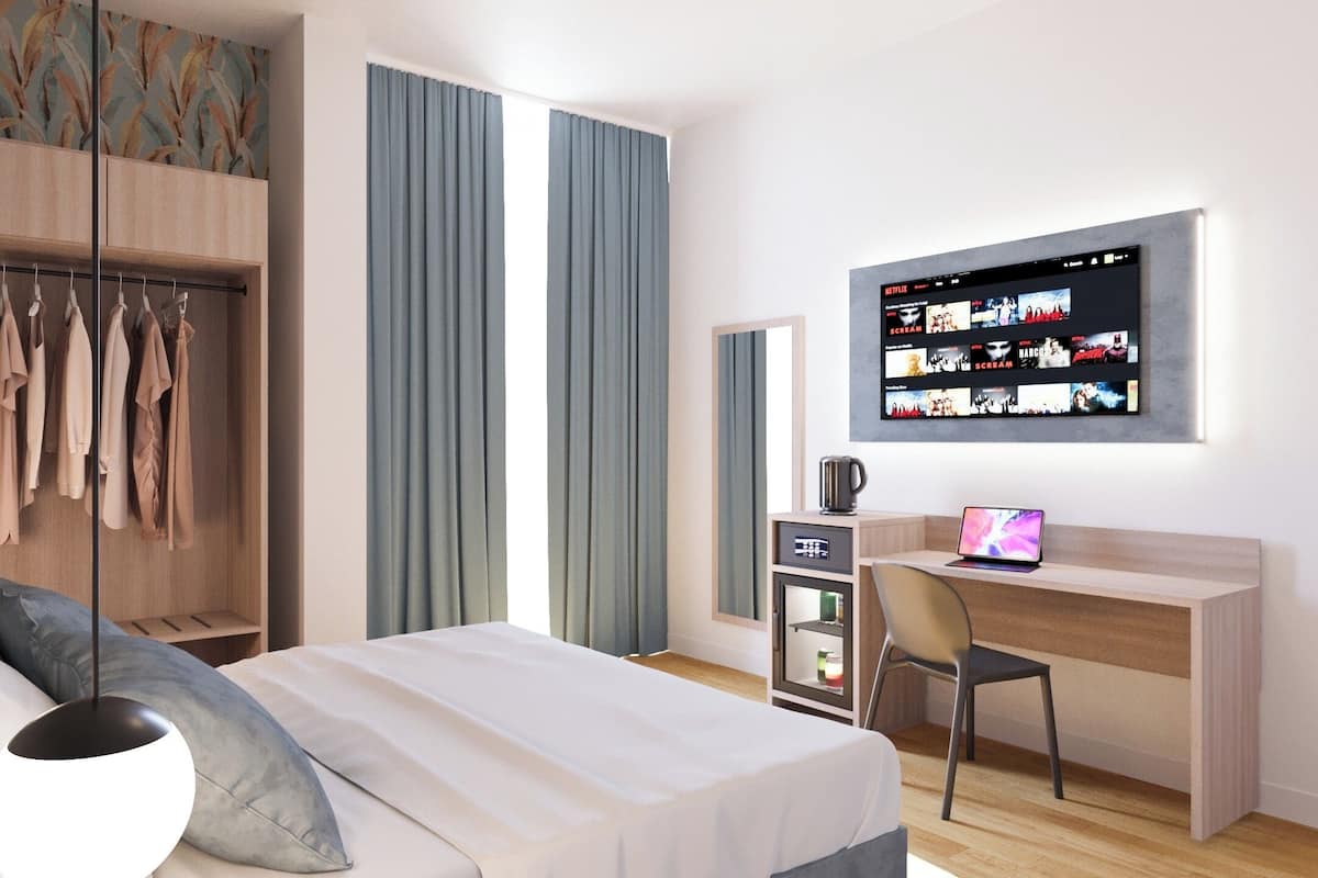 Ein modernes Hotelzimmer mit Bett, Schreibtisch mit Laptop, Kleiderschrank und Wand-Fernseher, der eine Auswahl an Filmen zeigt.