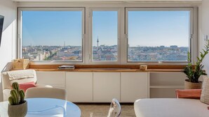 Una habitación con un amplio ventanal que ofrece vistas a la ciudad, un sofá blanco, una mesita con un cactus y un banco de madera con cojines.