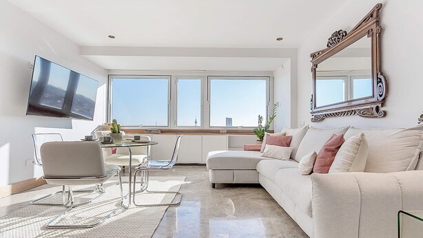 Una sala de estar moderna con un sofá, una mesa de comedor con sillas, un televisor de pantalla plana montado en la pared y ventanas amplias con vistas a la ciudad.